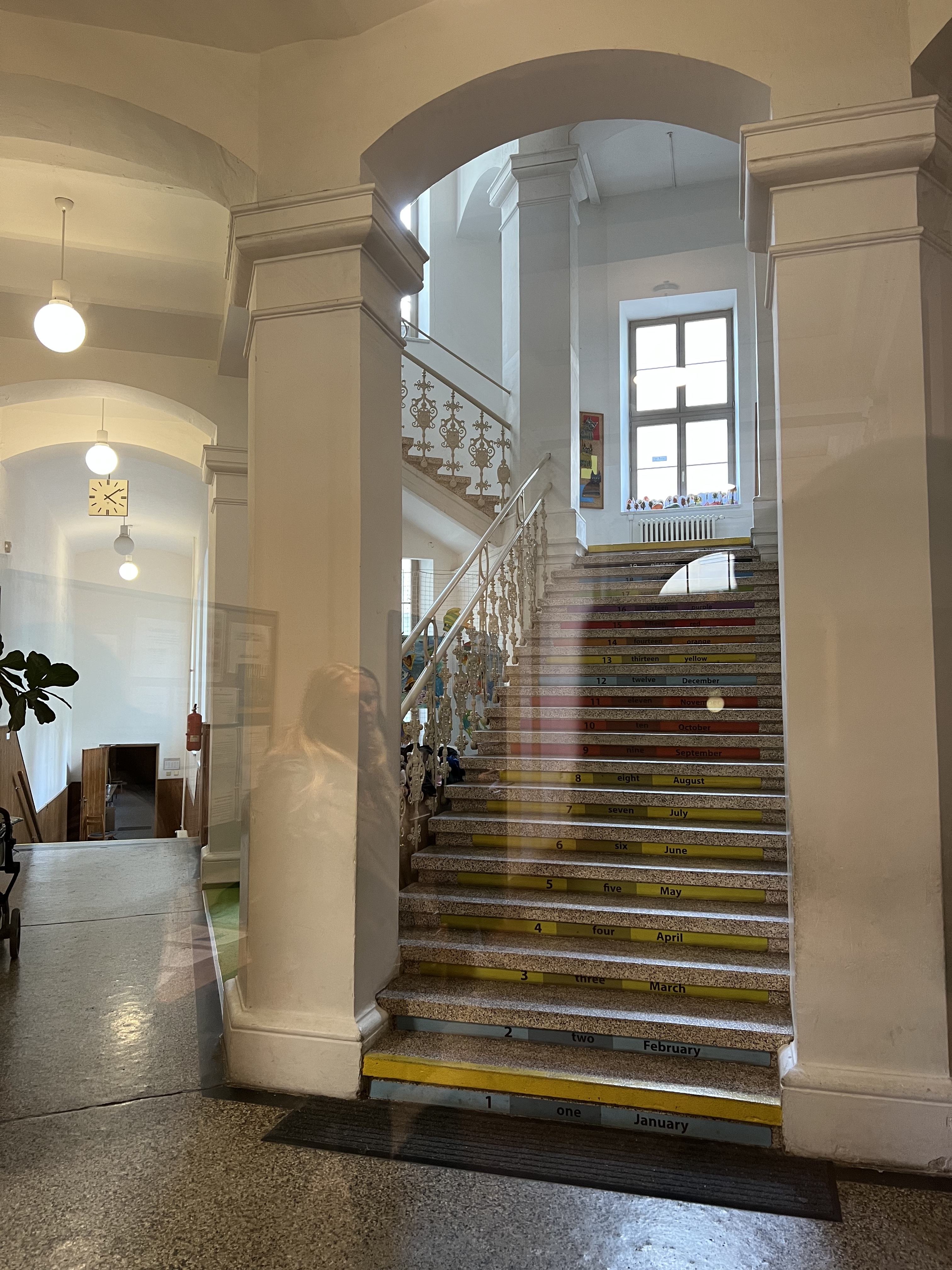 A large T-shaped stairwell rises between columns to a landing about one and a half floors above ground; reflections of the space behind the photographer appear in the glass door through which the photo was taken.